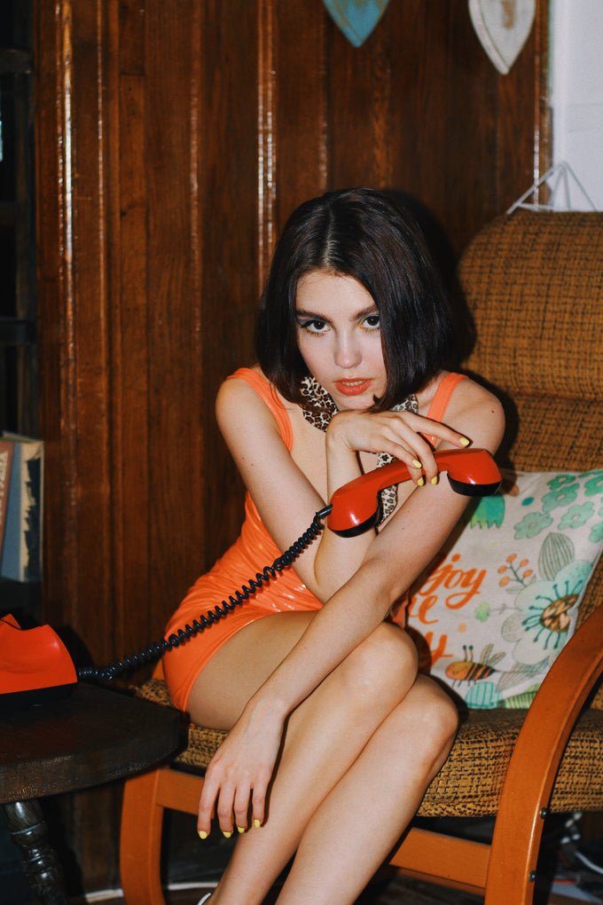  Caucasian woman in bodycon orange dress sitting in a retro 70s chair and holding a red phone